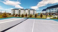 Located at 9 Monticello Dr in Brewster Village, NY, this outdoor recreational court facility offers a well-maintained, hard-surface court finished in a two-tone blue and gray color scheme with clearly marked playing lines. The court is fully equipped with a center net and is enclosed by sturdy black chain-link fencing, providing both safety and boundary definition. The facility appears designed to accommodate both tennis and pickleball, making it a versatile amenity for a variety of players and skill levels. Court-side lighting poles are in place, suggesting the potential for extended play beyond daylight hours. The surrounding landscape features neatly arranged evergreen shrubs and open green space, contributing to a clean and pleasant outdoor environment. Views from the court extend to the broader residential community, including multi-story residential buildings and a playground area visible in the background. The open-sky setting allows for abundant natural light throughout the day, and the surrounding scenery of trees and manicured grounds adds to the overall appeal. This court serves as a standout recreational feature within the Brewster Village community, offering residents a dedicated, purpose-built space for outdoor sport and activity.