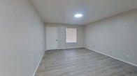 This single-floor unit presents a clean, modern aesthetic with a neutral color palette carried consistently throughout. Light gray walls, crisp white baseboards, and light-toned luxury vinyl plank flooring create a cohesive, well-maintained interior.

The main living area is open and functional, receiving natural light through a window fitted with blinds. A flush circular ceiling light provides overhead illumination, while a dedicated entry door with a standard lock offers direct access. An adjacent open area extends the usable living space, separated by a partial wall that defines distinct zones without closing off the floor plan.

The bedroom is a well-proportioned, private room featuring a ceiling fan with an integrated light fixture and a window that brings in natural light. Clean walls and matching flooring maintain the consistent style of the unit.

The kitchen is notably updated, featuring white shaker-style cabinetry with matte black hardware, granite-look countertops, a white subway tile backsplash, a matte black pull-down faucet, and stainless steel appliances including a refrigerator. A range hood is also present.

The bathroom is finished with a white vanity, round mirror, matte black accents, a combination tub and shower, and a toilet. Light sconce fixtures add a modern touch. The space as a whole is well-presented and move-in ready.