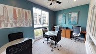 This single-floor business center offers a thoughtfully designed workspace that blends functionality with a coastal-inspired aesthetic. The room is defined by bold teal accent walls paired with neutral gray tones, creating a vibrant yet focused atmosphere well-suited for productivity.

Large sliding windows allow abundant natural light to fill the space, offering views of the surrounding exterior and greenery. A ceiling fan provides additional comfort overhead, complementing the recessed lighting throughout.

The layout accommodates both collaborative and individual work styles. A central round table with ergonomic rolling chairs invites group discussions or casual meetings, while a dedicated workstation along the wall is outfitted with computers, a printer, and ample desk surface for task-focused work.

Decorative framed artwork enhances the walls, including a surf-themed print and geometric coastal-inspired canvases, reinforcing the relaxed yet professional character of the room. Carpeted flooring adds warmth underfoot and contributes to a quiet, comfortable environment.

Overall, this business center strikes an inviting balance between a polished professional workspace and a welcoming, visually engaging design — making it an ideal setting for residents or guests seeking a productive and comfortable place to work.