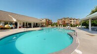 Located at 200 Governors Ct in Cartersville, Georgia, this outdoor amenity area serves as a premier recreational space within a residential community. The centerpiece is a generously sized freeform swimming pool with clear turquoise water, designed with gentle curves that give it a resort-like aesthetic. Adjacent to the main pool is a separate shallow wading area, ideal for those seeking a more relaxed aquatic experience. The expansive concrete sun deck surrounds both pools, providing ample space for lounge chairs and patio umbrellas, allowing residents to comfortably enjoy the outdoors. A covered pavilion with a pitched roof anchors one side of the deck, offering a shaded gathering space with additional seating and tables — perfect for socializing or escaping the Georgia sun. The entire pool area is enclosed by a wrought iron fence, providing both security and an open, airy feel. Tall, mature pine trees line the perimeter, creating a natural green backdrop that enhances the sense of seclusion and tranquility. The multi-story residential buildings visible in the background reflect a classic architectural style with warm earth tones and balconies, adding a cohesive community character to the setting. This well-appointed outdoor space combines functional design with inviting aesthetics for year-round enjoyment.