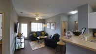 This single-floor unit showcases an open-concept layout that seamlessly connects the living area and kitchen. The living room is anchored by plush dark leather sofas accented with vibrant yellow-green and teal throw pillows, arranged around a glass-top coffee table atop a circular patterned area rug. A ceiling fan with integrated lighting fixture sits centrally overhead, providing both airflow and ambient illumination. Large paneled windows and a glass door along one wall invite generous natural light into the space, framed by floor-length curtains. The walls are finished in a cool gray tone, complementing the dark furnishings and creating a modern, cohesive aesthetic. Framed artwork decorates the walls, adding visual interest. The kitchen area, partially visible behind a breakfast bar with bar stool seating, features light-toned countertops and offers a convenient pass-through feel that enhances the openness of the floor plan. A hallway visible in the background leads to additional rooms, suggesting a thoughtful and functional layout. The carpeted flooring throughout the living space adds warmth and comfort to the overall atmosphere.