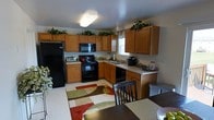 This two-story home offers a practical and comfortable layout designed for everyday living. The kitchen serves as a central hub, featuring warm honey-toned wood cabinetry that extends to the upper walls, providing generous storage throughout. Black appliances, including a full-size refrigerator, electric range, built-in microwave, and dishwasher, lend a cohesive, modern contrast to the cabinetry. A double-basin sink sits beneath a window, allowing natural light to filter into the space. The countertops offer ample workspace, and additional small appliances are neatly arranged along the backsplash. Decorative greenery displayed atop the upper cabinets adds a touch of warmth and personality to the room. The kitchen flows naturally into an adjacent dining area, where a dark-stained dining table and chairs are positioned over a colorful geometric area rug, adding visual interest to the space. The layout encourages an open, connected feel between the cooking and dining areas. Flush ceiling lighting keeps the kitchen well-lit. The two-floor layout of the home provides additional living and sleeping space beyond what is visible in this area.