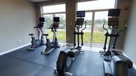 This fitness room offers a bright and functional workout environment designed to support a range of cardio-focused exercise routines. The space is outfitted with multiple pieces of modern cardio equipment, including upright stationary bikes, elliptical cross-trainers with integrated display screens, and a rowing machine, providing users with diverse training options. The flooring consists of dark rubber gym tiles, offering both durability and cushioning underfoot during workouts.

A standout feature of this room is the expansive wall of large windows, which flood the space with natural light and provide open views onto a green roof terrace below, as well as the surrounding landscape of trees and neighboring structures. The windows extend nearly from floor to ceiling in sections, creating an airy, open atmosphere that enhances the workout experience. The neutral-toned walls complement the contemporary design of the equipment and keep the environment clean and focused.

Overall, the fitness room combines practical amenity with a pleasant visual connection to the outdoors, making it a well-considered space for daily exercise.