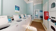 Located at 101 Breakers Dr in Myrtle Beach, South Carolina, this shared laundry facility is a practical and well-maintained single-floor space designed to serve its users efficiently. The interior is finished with soft aqua-toned walls and light ceramic tile flooring, creating a clean and pleasant atmosphere enhanced by natural light streaming through arched, blinds-fitted windows.

The main area houses a generous row of top-load washing machines along one wall, complemented by a separate section featuring stacked dryer units, providing ample capacity for multiple users simultaneously. Each machine is equipped with a digital payment interface, supporting app-based transactions for added convenience. A red vending or payment kiosk is also present near the entrance for additional payment options.

A ceiling fan keeps the space well-ventilated, while a wall-mounted television offers entertainment during laundry cycles. Entry and exit is provided through a glass-panel door, allowing natural light to filter through. A folding counter and waste receptacle near the entrance offer practical utility for users.

Rugs placed along the main walking paths add comfort underfoot and help define the circulation areas within the space. Posted signage throughout the facility provides clear instructions on machine operation and facility rules. Overall, this laundry facility is thoughtfully appointed to deliver a functional, user-friendly experience.