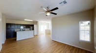 This two-floor residence offers a well-designed layout that balances comfort and functionality. The main living area is open and airy, featuring warm hardwood-style flooring that extends throughout the space, creating a cohesive and inviting atmosphere. A ceiling fan with a built-in light fixture anchors the living room, providing both airflow and ambient lighting.

The kitchen is seamlessly integrated into the open-concept floor plan and is equipped with stainless steel appliances, including a refrigerator and range. Dark cabinetry provides a striking contrast against the light-toned countertops and walls, lending the kitchen a modern, polished look. A kitchen island or breakfast bar adds additional prep and seating space.

Adjacent to the kitchen and living area, a naturally lit room — likely a dining space or secondary sitting area — benefits from a window that draws in natural light, further enhancing the bright and welcoming character of the main floor. A staircase provides access to the second floor, which offers additional living or sleeping quarters.

Throughout the home, neutral wall tones, clean white trim, and consistent flooring create a unified aesthetic that is both practical and visually appealing. The thoughtful layout and modern finishes make this a functional and comfortable multi-level residence.