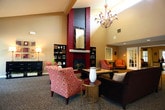 Located at 260 Marcella Rd in Hampton, Virginia, this single-floor community space is thoughtfully designed to offer both comfort and functionality. The layout encompasses several distinct areas, each serving a specific purpose while maintaining a cohesive aesthetic throughout.

The main lounge area features plush carpeting and is anchored by a comfortable dark upholstered sofa, accented with decorative throw pillows. Large windows dressed in elegant layered curtains flood the room with natural light, while French doors provide direct access to an outdoor area. Tall indoor plants add a lively, welcoming touch to the space.

Adjacent to the lounge is a dedicated business center, finished with large-format tile flooring and surrounded by expansive windows that offer abundant natural light and views of the outdoors. The area is equipped with multiple workstations and complemented by a round meeting table with upholstered red chairs, making it ideal for both individual work and group collaboration.

The kitchen is efficiently configured in an L-shape layout, featuring dark espresso cabinetry, light countertops, a stainless steel double-basin sink, and a dishwasher. A window above the sink provides ventilation and natural light.

A carpeted bedroom or private room rounds out the space, showcasing warm neutral tones, built-in shelving, and ample room for furnishings. Together, these areas create a versatile, inviting environment suited for residential community use.