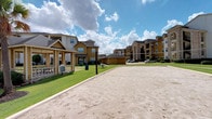 This community amenity center serves as the social hub of a well-appointed residential complex, combining thoughtful architectural design with an impressive array of outdoor recreational features. The building's exterior showcases a refined blend of natural stone columns and crisp white horizontal siding, creating a classic yet inviting aesthetic. Large multi-pane windows with arched transoms line the façade, flooding the interior with natural light and offering views of the surrounding grounds. A covered walkway with sturdy stone pillars runs along the exterior, providing a shaded passage to the entrance.

At the heart of the outdoor amenities is a generously sized resort-style swimming pool, its freeform shape set against a spacious concrete deck surrounded by towering palm trees that lend a tropical ambiance. A decorative water feature with stone accents adds an elegant focal point to the pool area. Adjacent to the building, a regulation sand volleyball court provides a dedicated space for active recreation, bordered by manicured green lawn. The broader community, visible in the background, features multi-story residential buildings with consistent architectural styling, creating a cohesive and attractive neighborhood setting beneath open skies.
