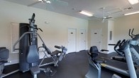 This single-floor fitness facility offers a functional and practical workout environment designed to accommodate a variety of exercise needs. The room features dark rubber flooring, which provides cushioning and durability suited for gym equipment and physical activity. Multiple ceiling fans are installed throughout the space, ensuring consistent air circulation during workouts, complemented by fluorescent strip lighting that brightens the area evenly.

The room is furnished with several pieces of exercise equipment, including a multi-function weight machine and a seated rowing or resistance machine, providing options for both strength training and cardiovascular exercise. The walls are painted in a neutral off-white tone, contributing to a clean and open atmosphere.

Along the back wall, three white panel doors with signage lead to additional rooms, likely changing areas, restrooms, or storage facilities, enhancing the overall utility of the space. The layout allows for comfortable movement between equipment. The ceiling height adds to the sense of openness, making the fitness area feel spacious and well-ventilated — an inviting environment for regular exercise routines.