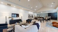 This well-appointed single-floor space offers an open, flowing layout that seamlessly integrates a lounge, dining, and kitchen area into one cohesive environment. The interior is defined by light wide-plank hardwood flooring, vaulted shiplap ceilings with recessed lighting, and crisp white walls that together create a bright and airy ambiance throughout.

The lounge area is anchored by a plush sectional sofa and contemporary armchairs in navy and neutral tones, complemented by a wall-mounted television and a dark-finished media console. Bold abstract artwork adorns the walls, adding visual interest and a modern sensibility to the space.

The dining area accommodates multiple seating arrangements, featuring round marble-top tables paired with rattan and dark wood chairs — ideal for social gatherings. A dramatic gallery wall of large-scale botanical prints provides a striking focal point nearby.

The kitchen is equipped with a substantial island topped with light stone, dark matte cabinetry reaching floor to ceiling, stainless steel appliances, and a textured stone tile accent wall that adds depth and character. Marble tile flooring transitions into this zone, distinguishing it subtly from the main living areas.

French doors with multi-pane glazing open to an outdoor deck, inviting natural light deep into the interior and offering views of the surrounding exterior. The overall design strikes a balance between refined contemporary style and welcoming functionality.
