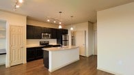 This single-floor space showcases a well-designed open-concept kitchen that serves as the centerpiece of the layout. The kitchen is outfitted with rich, dark cabinetry that contrasts sharply against light-toned walls and granite countertops, creating a bold, contemporary aesthetic. A central island with a built-in sink and matching granite surface provides additional workspace and doubles as a casual gathering spot. Stainless steel appliances, including a refrigerator, range, and microwave, complement the modern design scheme. Overhead, a combination of track lighting and elegant pendant lights ensures ample illumination throughout the cooking and dining area. The warm hardwood flooring extends seamlessly across the open space, grounding the design and adding a sense of continuity. The open layout connects the kitchen to the adjacent living area, enhancing the sense of spaciousness. Neutral wall tones throughout keep the atmosphere clean and versatile. Additional doorways visible from the main area suggest access to further rooms, contributing to a functional and well-organized floor plan overall.