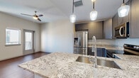 This well-appointed 2-bedroom, 1-bathroom residence offers a thoughtfully designed open-concept layout that seamlessly connects the kitchen, dining, and living areas into one cohesive space. The kitchen is a standout feature, showcasing gray shaker-style cabinetry, granite countertops, a peninsula island with a built-in sink, pendant lighting, and a full suite of stainless steel appliances including a French door refrigerator, range, microwave, and dishwasher. The kitchen transitions into the living area, which is bathed in natural light from large windows and a glass door leading to a private balcony. Rich hardwood flooring flows throughout the main living areas and bedrooms, lending warmth and continuity. The primary bedroom is generously sized, featuring a ceiling fan, large windows with outdoor views, and ample natural light. The second bedroom includes a built-in closet with shelving for convenient storage. Four closets throughout the unit ensure excellent storage capacity. The bathroom is elegantly finished with marble-style tile, a vanity with an illuminated mirror, and a soaking tub with a tiled surround. A dedicated laundry area and hallway complete this well-organized, modern home.