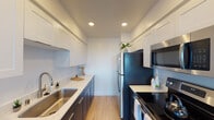 This well-appointed single-floor residence offers a clean, modern aesthetic throughout, with thoughtful updates and an abundance of natural light. The galley-style kitchen is a standout feature, showcasing two-tone cabinetry with crisp white upper cabinets and deep charcoal lower cabinets, complemented by bright white countertops and stainless steel appliances including a refrigerator, electric range, and built-in microwave. Light wood-look flooring runs underfoot, adding warmth to the space. The open living area is generously proportioned, featuring the same light wood flooring, a ceiling fan, and large floor-to-ceiling windows that frame sweeping views of palm-lined streets and the surrounding cityscape, flooding the room with natural light. The bedrooms are finished with soft, neutral-toned carpet and ceiling fans, providing a comfortable retreat. One bedroom benefits from a mirrored sliding closet and direct access to an outdoor-facing window with panoramic views. The bathroom is elegantly appointed with a wide undermount sink, marble-style quartz countertop, dark wood cabinetry, and a large backlit mirror. Baseboard heating is present throughout, and the overall palette of whites and soft neutrals lends the entire space a clean, airy ambiance.