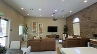 This single-story space is defined by its open, airy living area that immediately draws attention with its generous ceiling height and abundance of recessed lighting. A ceiling fan anchors the center of the room, contributing to both the aesthetic and functional comfort of the space. The living area is designed around a wall-mounted flat-screen television set atop a warm wood media console, flanked by framed abstract artwork that adds a touch of contemporary character to the neutral, beige-toned walls. Comfortable brown upholstered seating is thoughtfully arranged to create an inviting gathering space, complemented by decorative throw pillows and tall potted plants that bring organic warmth to the interior. A standout architectural feature is the exposed brick accent wall on one side of the room, adding texture and a rustic contrast to the otherwise modern finishes. An arched window on the brick wall allows natural light to filter in, enhancing the bright and welcoming atmosphere. Light-toned tile flooring runs throughout, contributing to the clean, cohesive look of the space. Overall, the design balances contemporary styling with classic elements, resulting in a polished and comfortable environment.