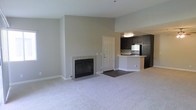 This single-floor residence offers a clean, open-concept layout that seamlessly connects the living area to the kitchen, creating a functional and comfortable living environment. The living room is anchored by a classic fireplace set against a neutral-toned wall, providing a focal point for the space. Soft, light-colored carpeting extends throughout the living area, contributing to a warm and inviting atmosphere. Vaulted ceilings enhance the sense of openness, allowing the space to feel airy and expansive. The transition from the carpeted living area to the kitchen is marked by a shift to hardwood-style flooring, clearly defining each zone. The kitchen is equipped with dark espresso-toned cabinetry that provides generous storage, complemented by stainless steel appliances including a refrigerator and range. A peninsula-style breakfast bar with a light countertop offers casual dining and additional workspace. The neutral gray-beige wall palette throughout creates a cohesive, modern aesthetic that pairs well with both light and dark furnishings. An interior door off the living area provides convenient access to additional areas of the unit. Overall, the space is designed with practicality and comfort in mind, offering a welcoming environment suitable for everyday living.