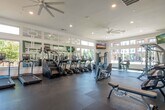 This fitness center is a thoughtfully designed, single-level space that offers a comprehensive workout environment. The layout is divided into distinct functional zones, each equipped to support a variety of exercise routines.

The cardio area is the most prominent section, featuring multiple treadmills, stair climbers, ellipticals, and a rowing machine arranged to maximize floor space. Large windows line the walls, flooding the area with natural light and providing outdoor views of surrounding greenery and the adjacent pool courtyard. A wall-mounted flat-screen TV and a ceiling fan overhead enhance comfort during workouts.

A decorative geometric mirror partition elegantly separates the cardio zone from the strength training area, adding visual depth while reflecting light throughout the space. The strength section includes a cable machine, a multi-functional rack with barbells, a weight bench, and a full dumbbell rack, catering to a wide range of resistance training needs.

A separate area near the entrance provides additional open floor space with a weight bench, a wall-mounted TV, and a small refreshment station with a beverage cooler. Glass doors lead directly outdoors. Recessed lighting, dark rubber flooring, and neutral-toned walls complete the clean, modern aesthetic throughout.