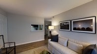 This single-story space showcases a thoughtfully designed living area that serves as the central hub of the home. The living room features warm hardwood-style flooring that runs throughout, complemented by smooth, light gray walls that create a clean and contemporary backdrop. A pass-through window connects the living area to the kitchen, offering a glimpse of dark cabinetry and providing an open, communicative feel between the two spaces. The kitchen is visible through the cutout, suggesting a functional and well-appointed layout. Framed black-and-white artwork adorns the main wall of the living room, adding a refined, gallery-like touch to the space. A hallway extends toward the back of the home, hinting at additional rooms beyond. The ceilings are smooth and of a comfortable height, contributing to the airy atmosphere. Natural and artificial lighting work in tandem to keep the space feeling bright and welcoming. Overall, the design strikes a balance between modern minimalism and comfortable livability, making it a practical yet stylish environment.