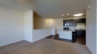This single-floor space showcases an open-concept layout that seamlessly connects the living area to the kitchen. The living area is finished with plush, neutral-toned carpet, creating a warm and comfortable atmosphere, while the kitchen and adjoining spaces transition to rich, dark hardwood flooring that adds a sharp visual contrast.

A defining architectural feature is the half-wall partition that subtly separates the living and kitchen zones while maintaining an open, airy feel throughout. Recessed lighting in the kitchen area provides focused illumination, complementing the flush ceiling light that brightens the living space.

The kitchen is well-appointed with dark espresso-toned cabinetry that extends to the upper walls, offering generous storage. A kitchen island with seating provides a functional and social centerpiece, while black appliances add a cohesive, contemporary aesthetic. A tiled backsplash adds texture and detail to the cooking area.

The neutral wall tones throughout the space contribute to a clean, understated palette, making it highly adaptable to a variety of interior styles. Overall, the layout balances practicality with modern design sensibility.