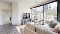 This two-bedroom, two-bathroom residence offers a contemporary single-level layout designed with clean lines and a neutral palette throughout. The open living area is anchored by expansive floor-to-ceiling windows that frame urban cityscape views, flooding the space with natural light and creating a striking visual connection to the surroundings. Hardwood-style flooring runs through the main living area, complementing the bright, airy atmosphere.

The kitchen features white cabinetry, dark countertops, a subway tile backsplash, and stainless steel appliances, including a range and over-the-range microwave. A window above the counter adds additional natural light to the workspace.

Both bedrooms are carpeted and generously proportioned, each offering large windows with exterior views and ceiling fans. One bedroom includes a walk-in closet with built-in shelving, while the other benefits from a cozy, well-lit ambiance with wall-mounted lighting flanking the headboard wall.

The two bathrooms are finished with large-format tile flooring and white cabinetry. One bathroom features a glass-enclosed walk-in shower with marble-style tile surround, while the other includes an elongated vanity with dark countertops and integrated in-unit laundry storage. The overall aesthetic is cohesive, modern, and calm throughout.