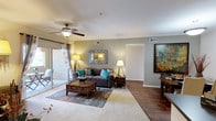 This single-floor residence offers a thoughtfully designed open layout that maximizes both comfort and functionality. The living area serves as the heart of the space, featuring a blend of plush carpeting and hardwood-style flooring that defines distinct zones within the room. Neutral gray walls provide a versatile backdrop, complemented by crisp white ceilings fitted with a ceiling fan for year-round comfort. Large sliding glass doors flood the living area with abundant natural light while offering direct access to an outdoor patio, extending the living space beyond its interior boundaries. The open flow between areas creates a spacious, airy ambiance well-suited for both relaxing and entertaining. Recessed air vents are integrated into the ceiling and walls, ensuring efficient climate control throughout. A wide doorway connects the living area to additional rooms, enhancing the sense of openness and ease of movement. The overall design balances practicality with an inviting warmth, making this a comfortable and versatile single-level living space.