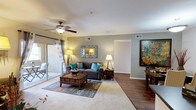 This single-floor residence offers a thoughtfully designed open layout that maximizes both comfort and functionality. The living area serves as the heart of the space, featuring a blend of plush carpeting and hardwood-style flooring that defines distinct zones within the room. Neutral gray walls provide a versatile backdrop, complemented by crisp white ceilings fitted with a ceiling fan for year-round comfort. Large sliding glass doors flood the living area with abundant natural light while offering direct access to an outdoor patio, extending the living space beyond its interior boundaries. The open flow between areas creates a spacious, airy ambiance well-suited for both relaxing and entertaining. Recessed air vents are integrated into the ceiling and walls, ensuring efficient climate control throughout. A wide doorway connects the living area to additional rooms, enhancing the sense of openness and ease of movement. The overall design balances practicality with an inviting warmth, making this a comfortable and versatile single-level living space.