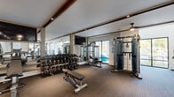 This fitness center offers a generously sized, single-level layout designed to accommodate a full range of workout activities. The space is divided into distinct functional zones, each thoughtfully outfitted with professional-grade equipment. The strength training area features an extensive dumbbell rack spanning a wide range of weights, multiple cable and functional training machines, a flat adjustable bench, and a large tire for functional fitness exercises. Full-length mirrors line the walls in this zone, amplifying the sense of space while allowing users to monitor their form. A wall-mounted television adds convenience overhead.

The cardio zone is positioned near floor-to-ceiling windows that frame views of lush outdoor greenery, creating a bright and inviting atmosphere. Treadmills, stationary bikes, and elliptical machines are arranged to take full advantage of the natural light and outdoor sightlines. Soaring ceilings in this section further enhance the open, airy feel.

Warm wood beam accents run across the ceiling throughout, complemented by recessed lighting and ceiling fans that contribute to a polished yet functional aesthetic. Durable carpeted flooring covers the entire space, providing comfort and traction. A small storage station with cubbies and yoga mats is also available, completing this well-rounded fitness facility.