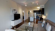 This single-floor residence showcases a thoughtfully designed open-concept layout that connects the living area and kitchen into one cohesive, functional space. The living room is anchored by a low-profile white media console supporting a flat-screen television, complemented by a patterned area rug, a rectangular coffee table, and comfortable seating that creates an inviting atmosphere. Track lighting runs along the ceiling, providing adjustable illumination across the space, while a pendant light fixture adds a decorative touch above the kitchen island area.

The kitchen is finished with sleek dark cabinetry that contrasts sharply against light-colored walls and countertops, giving the space a contemporary, polished look. Stainless steel appliances, including a refrigerator and range, are integrated seamlessly into the cabinetry. A kitchen island with bar stool seating offers a casual dining option and serves as a natural divider between the culinary and living spaces. Rich, dark hardwood flooring flows consistently throughout, adding warmth and cohesion. A hallway visible in the background suggests additional rooms beyond the main living area, completing this well-appointed residence.