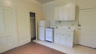 This single-floor space centers around a compact but functional kitchen area. The kitchen is equipped with essential white appliances, including a full-sized refrigerator and a gas range, alongside a single-basin sink set into a white cabinet base. Upper white cabinetry provides overhead storage, while lower cabinets beneath the sink offer additional space for organization. The flooring in the kitchen area features light-toned vinyl tile, which transitions to a darker solid flooring in the adjacent area, visually distinguishing the kitchen zone from the rest of the space. The walls are painted in a neutral off-white tone, contributing to a clean, understated aesthetic throughout. An open doorway adjacent to the refrigerator leads to a closet, which includes a hanging rod, offering practical storage solutions. The layout is straightforward and efficient, making practical use of the available space. Natural light appears to supplement the interior, keeping the space feeling open despite its modest footprint. Overall, the unit offers a no-frills, utilitarian design suited for everyday living.