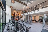 Located at 401 N Main St in Santa Ana, CA, this contemporary fitness center offers a well-appointed, spacious workout environment spread across two levels. The main floor is anchored by soaring double-height ceilings, large-scale floor-to-ceiling windows that flood the space with natural light and provide views of the surrounding outdoor courtyard and urban landscape. Oversized ceiling fans and decorative pendant lighting complement the bright, open atmosphere.

The cardio zone features a row of Life Fitness treadmills and elliptical machines, each equipped with individual digital screens. Adjacent to this area, a striking large-format mural depicting athletic figures adds a bold, motivational focal point to the space. A multi-functional cable and resistance training rig, including battle ropes and suspension trainers, occupies another section of the main floor.

A dedicated free weights area includes a comprehensive dumbbell rack, adjustable benches, and plate-loaded barbells, all set against floor-to-ceiling mirrors that enhance the sense of depth. A separate functional training zone houses medicine balls, kettlebells, and stability balls alongside a ballet barre and mirrored wall. The entrance features a water bottle filling station and clean double-door access, while textured tile accent walls and neutral carpet flooring tie the design together throughout.