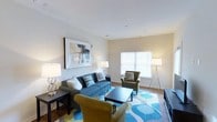 This single-floor space offers a clean, contemporary living environment designed for comfort and functionality. The living area serves as the centerpiece of the space, featuring smooth, light-toned walls and light hardwood-style flooring that contributes to an airy, open ambiance. A large window fitted with white horizontal blinds allows abundant natural light to fill the room, creating a warm and inviting atmosphere throughout the day. The living area is thoughtfully appointed, with ample space to accommodate a full seating arrangement, including a blue sofa and coordinating armchairs in complementary earth tones. A bold, geometric-patterned area rug in shades of blue, green, and white anchors the seating area and adds a vibrant focal point to the room. The walls are accented with tasteful artwork, adding a layer of visual interest without overwhelming the space. Recessed lighting and well-placed floor and table lamps provide layered illumination for both daytime and evening use. The overall design aesthetic is modern and cohesive, blending neutral architectural elements with colorful, carefully chosen furnishings to create a space that feels both polished and livable.