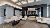 This single-floor amenity space is a study in contemporary luxury, seamlessly blending high-end materials with thoughtful design across several interconnected areas. The entrance lobby sets an immediate tone of sophistication, featuring large-format porcelain tile flooring, dramatic floor-to-ceiling marble-clad columns, and a sculptural backlit geometric wall panel as a focal point. A built-in banquette with tufted white upholstery, framed in rich dark wood millwork, offers an intimate seating alcove complemented by sleek pedestal tables.

The main lounge area is expansive and light-filled, with soaring coffered ceilings featuring recessed lighting that add architectural depth. A full glass curtain wall allows abundant natural light and provides open views of the surrounding outdoor space. Generous sectional sofas in neutral tones, layered rugs, and organic wood coffee tables create an inviting, resort-like atmosphere.

Adjacent to the lounge, a billiards and entertainment zone features a statement chandelier, wall-mounted media display, and stone accent walls. The gourmet kitchen is equally impressive, showcasing a long white waterfall island, dark wood cabinetry, stone-clad walls, and professional-grade stainless steel appliances, with elegant pendant lighting completing the refined aesthetic.