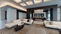 This single-floor amenity space is a study in contemporary luxury, seamlessly blending high-end materials with thoughtful design across several interconnected areas. The entrance lobby sets an immediate tone of sophistication, featuring large-format porcelain tile flooring, dramatic floor-to-ceiling marble-clad columns, and a sculptural backlit geometric wall panel as a focal point. A built-in banquette with tufted white upholstery, framed in rich dark wood millwork, offers an intimate seating alcove complemented by sleek pedestal tables.

The main lounge area is expansive and light-filled, with soaring coffered ceilings featuring recessed lighting that add architectural depth. A full glass curtain wall allows abundant natural light and provides open views of the surrounding outdoor space. Generous sectional sofas in neutral tones, layered rugs, and organic wood coffee tables create an inviting, resort-like atmosphere.

Adjacent to the lounge, a billiards and entertainment zone features a statement chandelier, wall-mounted media display, and stone accent walls. The gourmet kitchen is equally impressive, showcasing a long white waterfall island, dark wood cabinetry, stone-clad walls, and professional-grade stainless steel appliances, with elegant pendant lighting completing the refined aesthetic.