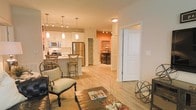 This thoughtfully designed single-floor residence offers an open-concept layout that seamlessly connects the living, dining, and kitchen areas, creating a spacious and airy feel throughout. The kitchen is a standout feature, equipped with stainless steel appliances including a refrigerator, microwave, and range, complemented by white cabinetry, a central island with bar seating, and pendant lighting that adds a modern touch. Light hardwood flooring runs throughout the main living areas, enhancing the warm and welcoming ambiance. The living area is generously sized and flows naturally into the rest of the home, making it ideal for both relaxing and entertaining. A striking burnt orange accent wall is visible beyond the main living space, adding a bold pop of color and character to the interior. Track lighting and flush-mount ceiling fixtures ensure the space is well-lit throughout. White panel doors with clean trim detail contribute to the crisp, polished aesthetic of the home. A secondary room, partially visible through an open doorway, hints at additional versatile living or dining space. Overall, the residence combines contemporary finishes with warm design choices to create a comfortable and functional living environment.