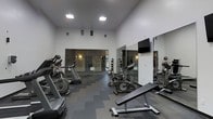 This fitness center offers a clean, modern environment designed to support a comprehensive workout experience. The space is defined by its impressive ceiling height, which lends an open and airy atmosphere, while recessed and flush-mounted overhead lighting ensures the area is well-lit throughout. The flooring consists of durable, dark rubber gym tiles that provide cushioning and traction during exercise. A key architectural feature is the full-length mirrored wall panels that line the perimeter, visually expanding the space and allowing users to monitor their form. Cardio equipment, including treadmills and stationary bikes, is arranged to maximize floor space and ease of movement between stations. A weight bench and additional strength training equipment are also present. A wall-mounted flat-screen television adds a modern amenity for entertainment during workouts. At the far end of the room, a glass-walled or mirrored partition separates an adjacent area, which appears to include additional equipment such as medicine balls and wall-mounted accessories. The overall ambiance is professional and functional, creating a focused environment well-suited for a range of fitness activities.