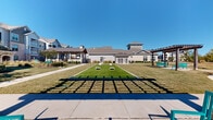 This expansive outdoor amenity space is thoughtfully designed to encourage recreation and relaxation within a contemporary residential community. At the heart of the grounds lies a dedicated bocce ball court, complete with a precisely maintained turf surface and cornhole boards nearby, offering residents multiple options for lawn games and casual outdoor entertainment. Two substantial wooden pergolas frame the activity areas, providing shaded seating with teal-painted benches beneath, ideal for spectating or simply enjoying the outdoors. Curved concrete pathways wind through the well-maintained lawn, connecting the various activity zones and creating a pleasant strolling environment. Carefully planned landscaping features young trees, ornamental grasses, and mulched planting beds that add texture and visual interest throughout the grounds. The perimeter of the property is defined by a sturdy wrought-iron fence with stone pillar accents, offering both security and a polished aesthetic. Detached garage units are conveniently situated nearby, complementing the residential buildings that rise in the background. The community structures reflect a modern farmhouse architectural style, incorporating board-and-batten siding, stone accents, and metal roofing. Under open Texas skies, this outdoor space delivers a vibrant, community-centered environment designed for active and leisurely use alike.
