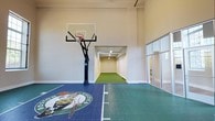 This exceptional indoor sports facility offers a remarkable combination of athletic amenities within a single, thoughtfully designed space. The centerpiece is a full-sized basketball court finished in a bold blue and green modular sport tile surface, prominently featuring the iconic Boston Celtics logo at center court. A regulation-height glass backboard and hoop system is mounted on a heavy-duty adjustable stand, providing an authentic on-court experience. The court is enclosed by tall, smooth walls painted in a clean neutral tone, lending an open and airy feel, while large floor-to-ceiling glass panels along one side allow generous natural light to filter into the space and offer views to an adjacent area. Directly connected to the basketball court is a dedicated indoor putting green, tucked into its own alcove and surfaced with lush artificial turf. Overhead track lighting ensures consistent illumination across the putting area. Together, these two distinct athletic zones create a versatile, private sports retreat that caters to a range of recreational pursuits. The overall design is clean and modern, balancing functionality with a polished, upscale aesthetic befitting a premier private sporting facility.