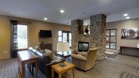This inviting single-floor space makes an immediate impression with its dramatic floor-to-ceiling stone fireplace, crafted from rounded river rock and serving as the undeniable focal point of the main lounge area. The fireplace features a glass-front firebox, allowing the warmth and glow of the fire to be enjoyed from multiple angles, and its broad stone mantel is adorned with tasteful decorative accents.

The lounge is thoughtfully furnished with a rich leather sofa, a plush upholstered armchair, and warm wood accent tables, creating a cozy and welcoming atmosphere. A table lamp with a classic white shade adds soft ambient lighting to complement the recessed ceiling lights overhead. A wall-mounted flat-screen television is positioned conveniently for comfortable viewing, accompanied by a dark wood dresser beneath it.

Natural light filters into the space through a window, brightening the neutral taupe walls and patterned area rug that anchors the seating arrangement. The overall ambiance strikes a balance between rustic lodge-inspired design and refined comfort, making the space feel both relaxed and well-appointed. The cohesive color palette of warm browns, tans, and grays ties the furnishings and architectural elements together seamlessly.