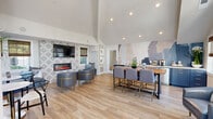 This thoughtfully designed single-floor space offers an inviting open-plan layout that maximizes its footprint through soaring vaulted ceilings and an abundance of natural light. Floor-to-ceiling windows and glass doors line the perimeter, flooding the interior with sunlight while providing views of the surrounding outdoor areas. Light-toned hardwood-style flooring runs throughout, enhancing the sense of openness and continuity.

The main lounge area serves as the focal point of the space, anchored by a sleek linear electric fireplace set within a bold geometric-patterned tile accent wall. A wall-mounted flat-screen TV sits above the fireplace, flanked by built-in banquette seating dressed with decorative pillows. Curved barrel chairs with gold-accented bases complement the contemporary aesthetic and encourage relaxed conversation. A live-edge wood communal table provides an additional gathering spot at the heart of the room.

The wet bar area features navy-blue cabinetry with brass hardware, white quartz countertops, an undermount sink, and stainless steel appliances, all set against a dramatic abstract watercolor mural that adds artistic depth. Woven bamboo Roman shades soften the natural light throughout. A large ceiling fan with elongated blades completes the airy ambiance, making this space as functional as it is visually striking.