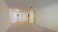 This single-floor unit presents a straightforward and functional layout centered around an open main living area. The space is finished with wall-to-wall carpeting in a neutral beige tone, complemented by cream-colored walls and white trim throughout, creating a clean and cohesive aesthetic.

The main room offers a generous open area suitable for a variety of furniture arrangements. A built-in closet with a sliding door provides convenient storage directly within the living space. The front entry door is centrally positioned and leads into the unit with ease.

Beyond the main area, a partitioned section visible from the living room suggests additional utility or storage space, with cabinetry hinting at a kitchen or kitchenette area nearby. The layout benefits from defined zones despite its open character, offering both functionality and organization.

Lighting is provided by flush-mounted ceiling fixtures, and a standard HVAC vent is centrally placed in the ceiling for climate control. White baseboards frame the perimeter of the room, adding a polished finish. Overall, the space maintains a simple, clean presentation well-suited for practical everyday living.