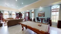 This well-appointed single-floor community space offers a variety of functional areas designed for both relaxation and recreation. The grand entry lobby sets an impressive tone with soaring vaulted ceilings, stacked stone columns, and an arched transom window above the main entrance that floods the space with natural light. Travertine tile flooring flows seamlessly throughout, lending a cohesive and polished feel to the entire facility.

The expansive game and lounge area serves as the heart of the space, featuring a full-size billiards table, a rich mahogany fireplace with built-in cabinetry, and a bar-height table and seating. Soft sage-green walls, crown molding, and multiple arched windows with floor-length drapery contribute to an inviting, refined ambiance. An adjacent seating area provides comfortable sofas and armchairs arranged around a built-in bookcase, ideal for quiet reading or casual conversation.

The kitchen is outfitted with dark cherry cabinetry, granite countertops, stainless steel appliances, and a window overlooking the outdoor area. A separate media or lounge room offers additional seating centered around a television, making it a versatile space for gatherings or quiet downtime. Throughout, recessed lighting and abundant natural light create a warm, welcoming atmosphere.