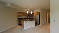 This single-floor residence showcases a thoughtfully designed open-concept layout that integrates the kitchen and living area into one cohesive space. The kitchen is a standout feature, equipped with stainless steel appliances including a full-size refrigerator, microwave, and dishwasher. Rich medium-toned wood cabinetry lines the walls, providing ample storage, while dark granite-style countertops add a sleek, contemporary contrast. A central island with a matching dark countertop serves as both a functional workspace and a casual gathering point. Overhead, a track lighting system runs along the ceiling, complemented by two elegant pendant lights that hang above the island, casting focused illumination over the prep area. The kitchen transitions smoothly into the adjoining living space, which features neutral-toned carpeting that extends throughout, contributing to a warm and comfortable atmosphere. The open sightlines between the kitchen and living area create a spacious, airy feel, making the layout well-suited for both everyday living and entertaining. Neutral wall tones and clean architectural lines throughout give the space a modern, understated aesthetic.