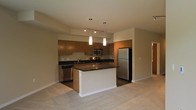 This single-floor residence showcases a thoughtfully designed open-concept layout that integrates the kitchen and living area into one cohesive space. The kitchen is a standout feature, equipped with stainless steel appliances including a full-size refrigerator, microwave, and dishwasher. Rich medium-toned wood cabinetry lines the walls, providing ample storage, while dark granite-style countertops add a sleek, contemporary contrast. A central island with a matching dark countertop serves as both a functional workspace and a casual gathering point. Overhead, a track lighting system runs along the ceiling, complemented by two elegant pendant lights that hang above the island, casting focused illumination over the prep area. The kitchen transitions smoothly into the adjoining living space, which features neutral-toned carpeting that extends throughout, contributing to a warm and comfortable atmosphere. The open sightlines between the kitchen and living area create a spacious, airy feel, making the layout well-suited for both everyday living and entertaining. Neutral wall tones and clean architectural lines throughout give the space a modern, understated aesthetic.