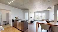 Located at 1140 Rue Wellington in Montréal's dynamic Griffintown district, this two-bedroom, one-bathroom unit showcases a contemporary urban aesthetic defined by striking exposed concrete ceilings and warm hardwood flooring throughout. The open-concept living, dining, and kitchen area creates a cohesive and functional living space. The modern kitchen is well-appointed with stainless steel appliances, including a range and refrigerator, sleek dark granite countertops, a black tile backsplash, high-gloss white upper cabinetry, and a peninsula with bar seating—ideal for casual dining or entertaining. The living room receives generous natural light through large sliding glass doors that open to an outdoor view, complemented by a bright, airy atmosphere. Both bedrooms feature the same consistent hardwood flooring and exposed concrete ceilings, lending an industrial-chic character. The primary bedroom offers a dedicated closet and ample space. The bathroom is cleanly finished with large-format tiles, a soaking tub with an overhead shower, and a white vanity. A dedicated in-unit laundry room with a stacked washer and dryer adds everyday convenience. Three closets provide excellent storage throughout the unit.