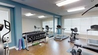 This fitness room is thoughtfully designed to accommodate a variety of workout styles within a compact, functional layout. The space features durable hardwood-style flooring that is both practical and easy to maintain, complemented by a textured ceiling with recessed fluorescent lighting that ensures the room is well-lit throughout. A large floor-to-ceiling mirror spans one wall, visually expanding the space while allowing users to monitor their form during exercise. The accent wall is painted in a deep teal tone, adding a dynamic and motivating atmosphere to the room. Natural light filters in through multiple windows fitted with white blinds, contributing to a bright and energizing environment. The room is equipped with cardio machines including a treadmill and a stationary recumbent bike, catering to endurance and low-impact training. A multi-tiered dumbbell rack holds a wide range of free weights, supported by additional accessories such as a stability ball, resistance balls, and a yoga mat. The overall ambiance is clean, functional, and focused, making it a well-rounded space for residents seeking convenient on-site fitness amenities.