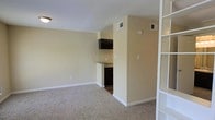 This single-floor unit offers a practical and efficiently designed open-concept layout. The main living area features wall-to-wall carpeting in a neutral tone, creating a warm and comfortable foundation throughout the space. Soft beige walls with clean white baseboards contribute to a bright, understated aesthetic that makes the space feel open and easy to personalize.

The kitchenette is partially partitioned from the living area by a low countertop divider, providing functional separation while maintaining visual connectivity. It is equipped with dark cabinetry, including an overhead cabinet and under-counter storage, along with a compact refrigerator. The countertop offers modest workspace for meal preparation.

A built-in shelving unit along one wall provides convenient open storage, adding both utility and a subtle architectural detail to the space. Overhead ventilation registers are present throughout, ensuring consistent climate control across the unit.

The layout is clean and straightforward, making it well-suited for efficient day-to-day living. Neutral finishes and a cohesive color palette create a move-in ready environment that is versatile and easy to furnish. Overall, the space delivers a functional and tidy living solution with thoughtful built-in features.