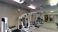 This fitness center offers a dedicated space for strength and resistance training, outfitted with multiple Paramount-brand cable and weight machines designed to target a full range of muscle groups. The machines are finished in white with black upholstered pads, lending the space a clean, professional aesthetic. A full-length mirror lines one wall, allowing users to monitor their form during exercise while also visually expanding the room. The flooring is a neutral-toned carpet, and the walls are painted in a warm taupe that creates a calm, focused atmosphere. Overhead, two ceiling fans work in tandem with bright panel and flush-mount lighting fixtures to maintain airflow and visibility throughout the space. A fire safety device and emergency lighting unit are mounted on the wall, reflecting attention to safety standards. The layout provides ample clearance between machines, allowing multiple users to exercise comfortably without congestion. Overall, the fitness center is thoughtfully designed to support an effective strength training routine in a clean, well-maintained, and practical environment.