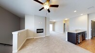 This single-floor space showcases an open-concept design that seamlessly connects the living area to adjacent spaces, creating a bright and airy atmosphere. The living room is anchored by a sleek, linear built-in fireplace set into a clean accent wall, serving as a focal point for the space. Overhead, a classic ceiling fan with a light fixture provides both comfort and illumination, complementing the neutral white ceiling above.

The flooring throughout the main living area consists of plush, light-toned carpeting that adds warmth underfoot. The walls are finished in a soft, neutral beige tone, lending the space a calm and cohesive aesthetic. Large baseboards frame the perimeter, adding a subtle architectural detail.

Toward the rear of the space, a doorway allows natural light to filter in, brightening the open layout. A partial view of a dark-toned kitchen island or bar area is visible to one side, hinting at an adjoining kitchen space. Additional doorways and transitional areas suggest multiple connected rooms, contributing to the overall spacious and functional flow of this well-designed, single-level home.
