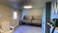 This single-floor utility space is purpose-built for pet washing and grooming. The room's most prominent features are two side-by-side stainless steel wash tubs, each equipped with flexible sprayer hose attachments and raised on sturdy metal frames, allowing for comfortable, elevated pet bathing. Protective metal splash guards between and behind the tubs help contain water during use.

The room is finished with light-toned ceramic tile flooring, which is both practical and easy to clean. Walls are painted in a neutral light green, contributing to a clean, functional atmosphere. A flush-mount circular ceiling light provides bright, even illumination throughout the space.

An exterior door with a window panel is positioned along one wall, offering natural light and direct outdoor access — a practical feature for moving pets in and out efficiently. A teal-painted interior door on the opposite side connects to the rest of the facility. A small waste bin near the exterior door adds to the room's practical layout. Overall, the space is designed with utility and cleanliness in mind, making it well-suited for regular pet care use.