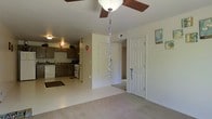 This single-story space offers an open, functional layout that connects the main living area to the kitchen. The living area features neutral beige carpeting and a ceiling fan with a built-in light fixture, contributing to a comfortable and well-lit environment. A paneled door leads to an adjacent room, adding to the flow of the floor plan.

The kitchen is visible from the living area and is equipped with dark wood cabinetry, a white range with oven, a refrigerator, and an under-cabinet range hood. Tile or vinyl flooring in the kitchen provides a practical contrast to the carpeted living space. The cabinetry offers ample storage, and the layout allows for efficient use of the cooking and preparation areas.

The neutral color palette of beige and white walls is consistent throughout, lending the space a clean and cohesive appearance. Overall, the open sightlines between rooms create a sense of connectivity, making the space feel accessible and easy to navigate. The straightforward design and practical finishes make it a functional and livable environment.