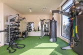 This fitness center is thoughtfully designed to support a wide range of workout routines across two distinct zones. The cardio area features light hardwood-style flooring and is lined with a row of treadmills, ellipticals, and stair climbers, complemented by a wall-mounted flat-screen TV. Large floor-to-ceiling windows flood this section with natural light and offer open views of the surrounding urban landscape.

Transitioning into the functional training zone, the flooring shifts to vibrant artificial turf, creating a dedicated space for dynamic and high-intensity workouts. This area is equipped with battle ropes, a heavy punching bag, medicine balls, resistance bands, a multi-use rack, and a cable machine. Stationary bikes and additional cardio equipment are also positioned throughout this space.

The strength training section includes a full dumbbell rack with a wide range of weights, a weight bench, a multi-station cable machine, and a selectorized strength machine. Full-length mirrors line the walls, enhancing the sense of openness and allowing users to monitor their form. Recessed lighting throughout provides consistent illumination across all zones. The overall layout is clean, open, and efficiently organized to accommodate a diverse range of fitness activities.