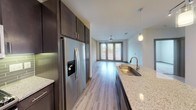 Located at Aura in Grand Prairie, Texas, this single-floor residence combines contemporary design with practical comfort throughout its thoughtfully laid-out spaces. The kitchen serves as a standout feature, showcasing rich dark cabinetry that extends to the ceiling for generous storage, a green subway tile backsplash, stainless steel appliances including a gas range, built-in microwave, and a French door refrigerator with an ice and water dispenser. Granite countertops and a large undermount sink complement the extended island with pendant lighting above, creating an ideal space for both cooking and entertaining. Light wood-style flooring flows seamlessly through the kitchen and living areas, contributing to a bright and cohesive aesthetic. The open living area benefits from natural light streaming through French doors, which also provide outdoor access. Track and ceiling-mounted lighting further enhance the space. The carpeted bedroom offers a comfortable retreat, featuring a ceiling fan, natural light from a window with blinds, and generous floor space. The bathroom is finished with matching dark cabinetry, granite countertops, an undermount sink, large mirror, wood-style flooring, and a tiled shower surround, maintaining the consistent modern style found throughout the residence.